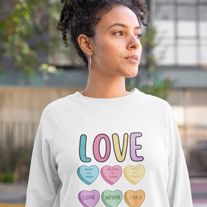 May include: White sweatshirt with a graphic of candy hearts that spell out "LOVE" and the text "1 Corinthians 13:7-8".
