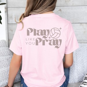 May include: Pink t-shirt with a volleyball graphic and the text "Play Like You Pray" in a retro font.
