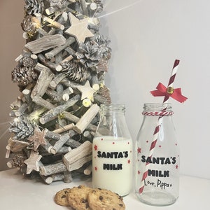 May include: Two glass milk bottles with "SANTA'S MILK" text and red star accents, one filled with milk. A red and white striped straw with a bow and bell sits in the empty bottle. Three chocolate chip cookies are in front.