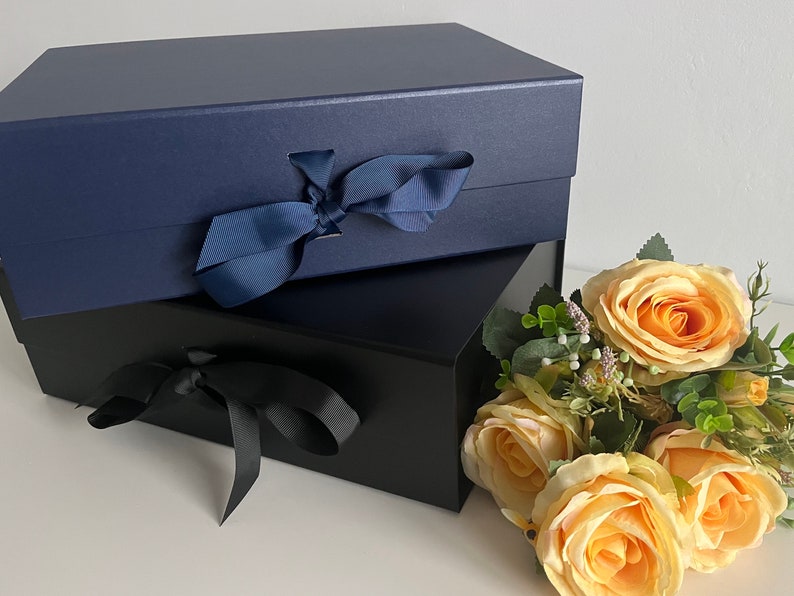 May include: Two gift boxes, one navy blue and one black, both tied with matching ribbon bows. The boxes are stacked on top of each other. A bouquet of yellow roses is in the foreground.