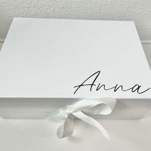 May include: White gift box with a white ribbon tied in a bow. The box has the name "Anna" written on the top in black cursive.