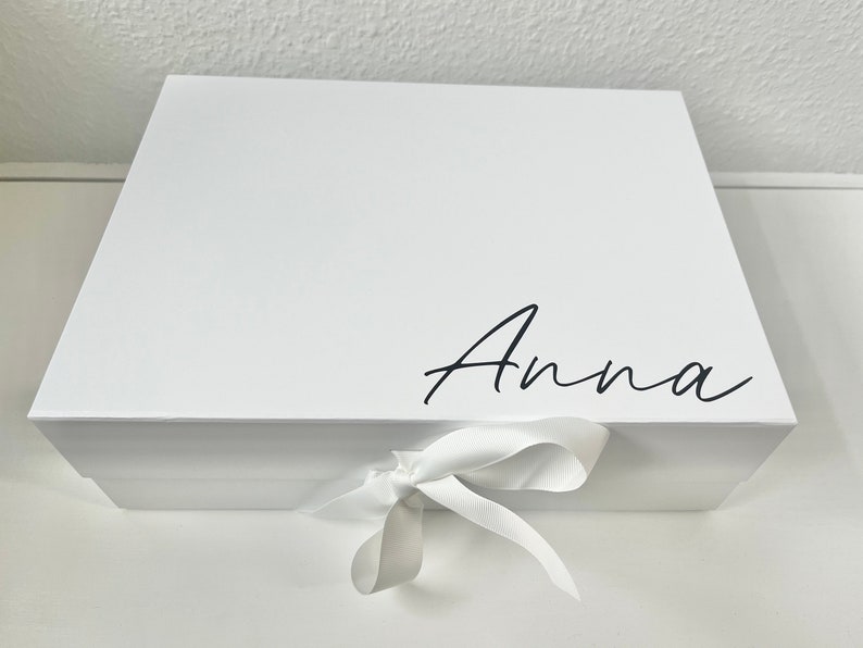 May include: White gift box with a white ribbon tied in a bow. The box has the name "Anna" written on the top in black cursive.