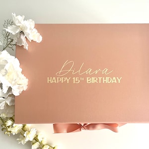May include: A blush pink gift box with a gold ribbon and the text "Dilara Happy 15th Birthday" on the lid.