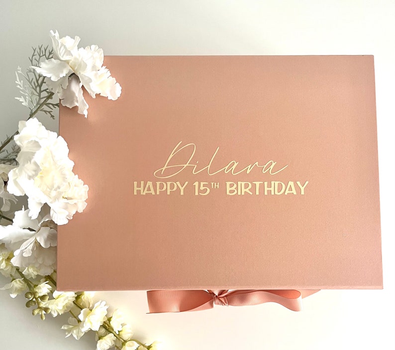 May include: A blush pink gift box with a gold ribbon and the text "Dilara Happy 15th Birthday" on the lid.