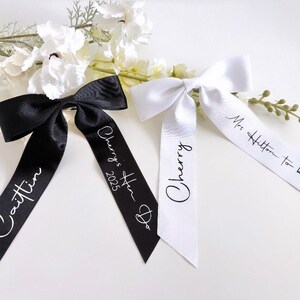 May include: Three satin ribbon bows, two black and one white, with personalized text. The black bows have the names "Caitlin" and "Cherry's Hen Do 2025" printed on them. The white bow says "Cherry" and "Mrs Hilton to be".