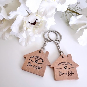 May include: A wooden house-shaped keychain with the text "Our First Home Est. 2025 Ben & Kate" engraved on it. The keychain is attached to a silver chain and two silver rings.