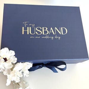 May include: A navy blue gift box tied with a matching ribbon. Gold text on the lid reads "To my HUSBAND on our wedding day". White flowers are in the lower left corner. The box is closed.