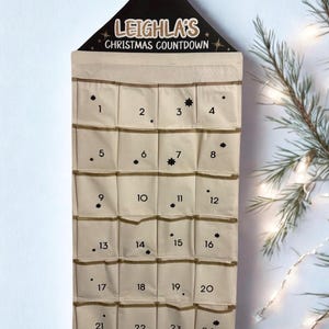 May include: A beige Christmas countdown calendar with 24 numbered pockets. The top section is black with the text "LEIGHLA'S CHRISTMAS COUNTDOWN". Each pocket is numbered and decorated with small star accents. A festive holiday decoration.