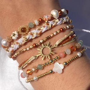 Op de afbeelding: Een verzameling armbanden met kralen in verschillende ontwerpen. De armbanden hebben parels, gouden kralen en bedels, waaronder een zon en een klavertjevier. Het kleurenpalet omvat wit, goud en pasteltinten, wat een bohemien esthetiek creëert.