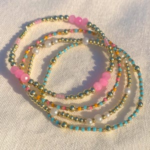 May include: A collection of beaded bracelets in various colors. The bracelets feature gold-colored beads, pink faceted beads, small turquoise, orange, and pink beads, and white pearl beads. The bracelets are arranged on a white surface.