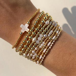以下が含まれることがあります： 金色のビーズブレスレットのコレクション。一部は白い真珠のアクセントがあり、1つは白い十字架のチャームが付いています。ブレスレットは手首に重ねられており、さまざまなデザインと質感が示されています。金色のビーズは光を反射しています。