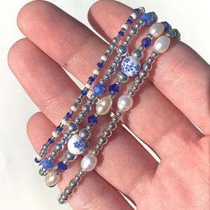 Pode incluir: Quatro pulseiras de contas em uma mão. As pulseiras apresentam contas prateadas, cristais azuis, contas brancas e pérolas. Duas pulseiras têm contas de cerâmica floral branca e azul. As pulseiras são empilhadas.