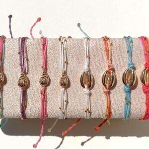 以下が含まれることがあります： カラフルな貝殻ブレスレットのコレクション。各ブレスレットは、金色の貝殻チャームと小さな金のビーズが特徴です。ブレスレットは、ピンク、紫、白、オレンジ、青など、さまざまな色の糸で作られています。