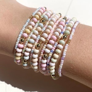 Könnte beinhalten: Ein Stapel pastellfarbener Perlenarmbänder mit goldenen Akzenten. Die Armbänder bestehen aus kleinen, runden Perlen und sind übereinander gelegt.