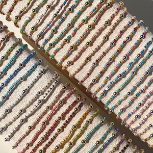 May include: A collection of colorful beaded bracelets displayed on a white surface. The bracelets feature a variety of colors, including blue, pink, green, and gold, with small beads and metallic accents. The bracelets are arranged in rows.