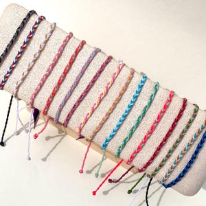 Puede incluir: Exhibición de pulseras de la amistad trenzadas en varios colores, incluyendo azul, rojo, rosa y verde. Las pulseras están dispuestas sobre un expositor cubierto de tela beige, mostrando sus patrones tejidos y hilos colgantes.