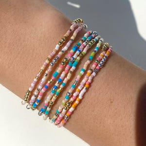 Puede incluir: Una colección de pulseras de cuentas en varios colores, incluyendo rosa, azul, amarillo y blanco. Las pulseras presentan pequeñas cuentas coloridas intercaladas con cuentas metálicas doradas y plateadas. Las pulseras están apiladas en una muñeca.