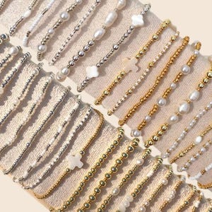 May include: An assortment of beaded bracelets in gold, silver, and white. Some bracelets feature pearls, while others have cross or flower-shaped charms. The bracelets are displayed on a beige fabric surface.