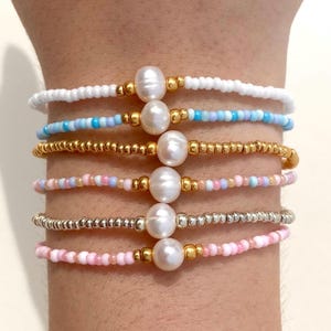May include: A collection of six beaded bracelets. Each bracelet features a different color scheme, including white, blue, gold, pink, and silver beads. Each bracelet has a large, oval-shaped pearl in the center, with gold-colored accents.