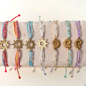 May include: A collection of colorful handmade bracelets. Each bracelet features a gold-colored charm, either a sun or a seashell, and is strung with a mix of colorful threads and small beads. The bracelets are displayed on a neutral fabric.