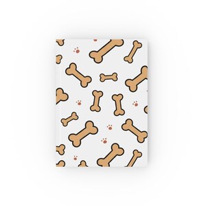 May include: A rectangular hardcover notebook featuring a repeating pattern of light brown dog bones and small reddish-brown paw prints against a solid white background. The notebook has a clean, flat design with a vertical spine on the left side.