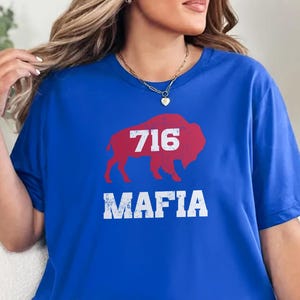 May include: A royal blue t-shirt featuring a red buffalo with the number "716" in white, and the word "MAFIA" in white below. The shirt is a short-sleeved crew neck.