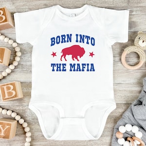 May include: White baby onesie with the text "BORN INTO THE MAFIA" in blue, a red buffalo graphic, and red stars. The onesie is short-sleeved and has a simple, classic design, suitable for infants.