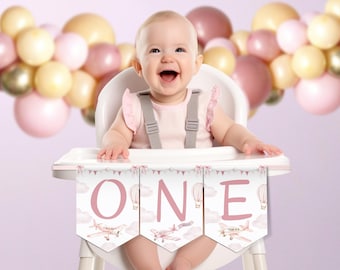 Könnte beinhalten: Ein Baby sitzt in einem Hochstuhl und lächelt, mit einem Banner mit der Aufschrift "ONE". Das Banner zeigt Illustrationen von Flugzeugen und Heißluftballons. Ballons in Rosa, Gold und Weiß sind im Hintergrund.