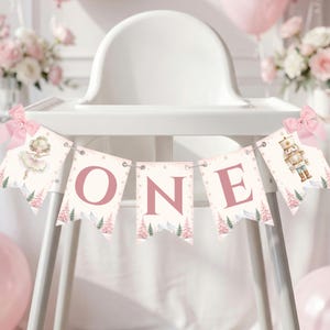 Pode incluir: Uma faixa decorativa com cinco bandeirolas e laços cor-de-rosa nas extremidades. As três bandeirolas centrais formam a palavra ONE em letras cor-de-rosa grandes. As bandeirolas exteriores exibem uma bailarina e um quebra-nozes.