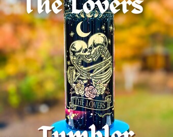 May include: A black and gold stainless steel tumbler with a design of two skeletons embracing, surrounded by roses and the text "The Lovers".
