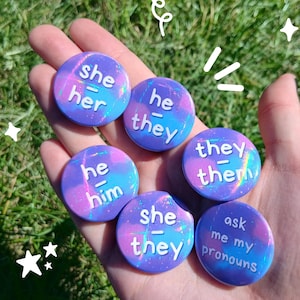 May include: Six round pronoun badges with a holographic purple, blue, and pink design. The badges display the pronouns "she/her", "he/they", "he/him", "they/them", "she/they", and "ask me my pronouns" in white text.