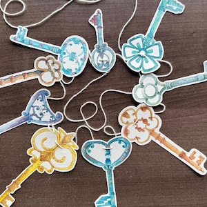 Puede incluir: Una colección de llaves decorativas de papel en varios colores, incluyendo azul, dorado y marrón, están dispuestas sobre una superficie marrón oscuro. Cada llave tiene un diseño único y está unida a una cuerda blanca.