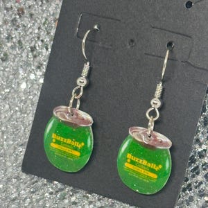 Könnte beinhalten: Ein Paar grüne Glitzer-Ohrringe in Form einer Dose Buzz Balls Cocktails. Die Ohrringe sind silberfarben und haben einen Hakenverschluss.