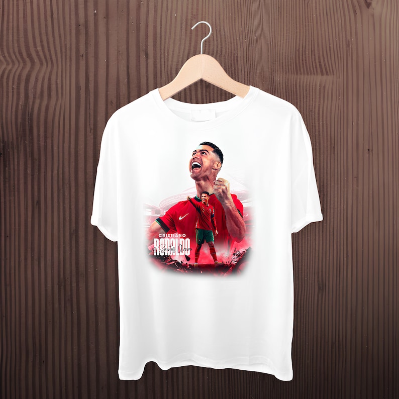 May include: White t-shirt with a graphic print of Cristiano Ronaldo celebrating a goal. The text "Cristiano Ronaldo" and his signature are printed on the shirt.