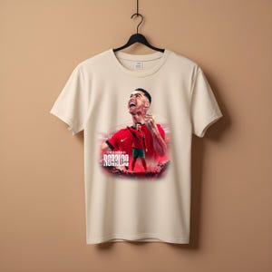 May include: White t-shirt with a graphic print of Cristiano Ronaldo in a red and green soccer uniform. The text "Cristiano Ronaldo" is printed below the image.