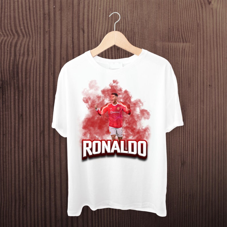 May include: White t-shirt with a red and white image of Cristiano Ronaldo in a red and white soccer uniform, with red smoke behind him. The text "RONALDO" is printed in bold, black letters below the image.