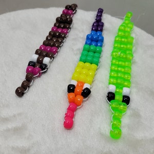 Könnte beinhalten: Drei perlenbesetzte, stiftförmige Accessoires. Eines ist braun, rosa und weiß. Das zweite ist regenbogenfarben. Das dritte ist hellgrün mit weißen und schwarzen Akzenten. Jedes ist auf einer weißen Schnur aufgefädelt.