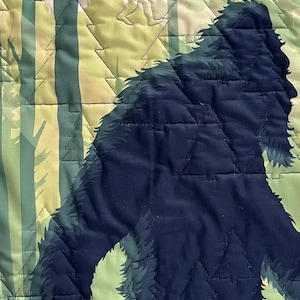 Puede incluir: Una tela acolchada azul oscuro con la silueta de una criatura grande y peluda caminando por un bosque. La criatura camina hacia el lado derecho de la imagen. El fondo es un patr&oacute;n de &aacute;rboles verde claro y marr&oacute;n.