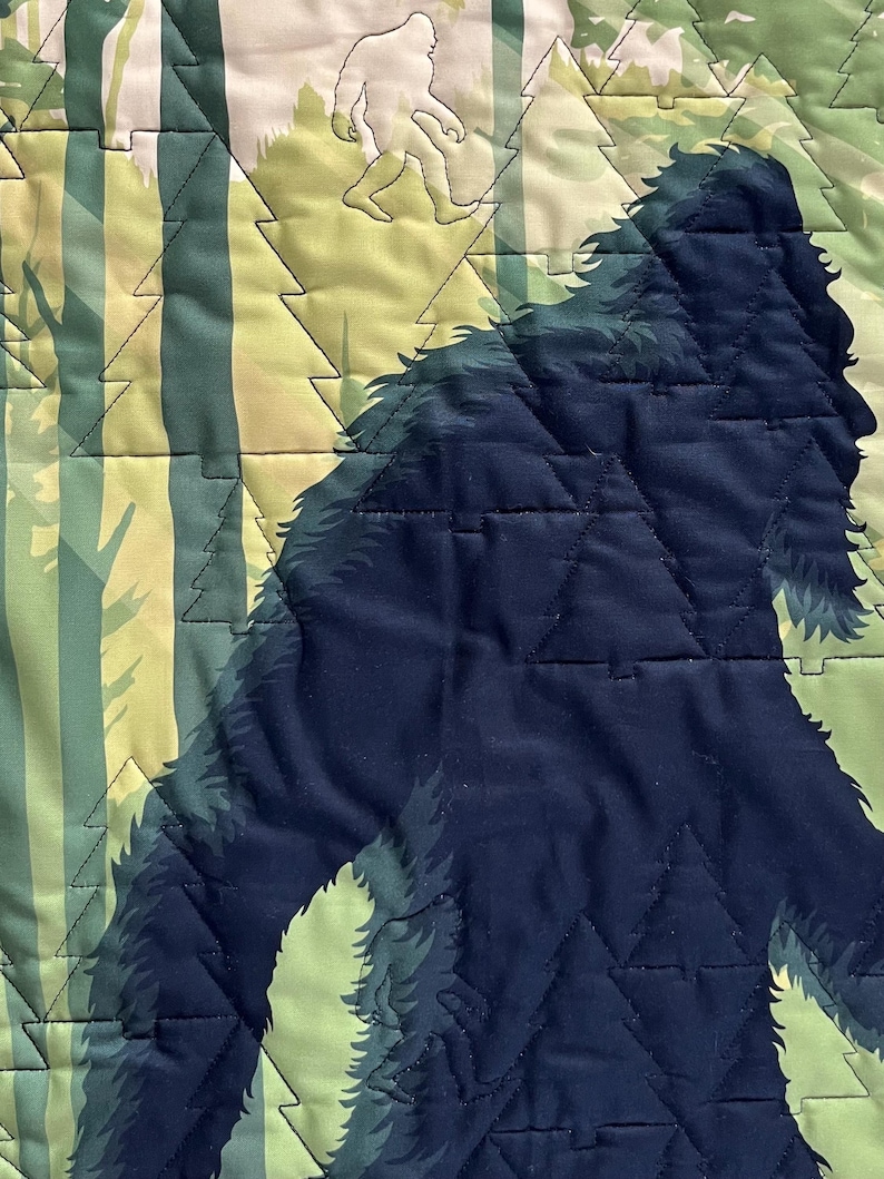 Puede incluir: Una tela acolchada azul oscuro con la silueta de una criatura grande y peluda caminando por un bosque. La criatura camina hacia el lado derecho de la imagen. El fondo es un patr&oacute;n de &aacute;rboles verde claro y marr&oacute;n.