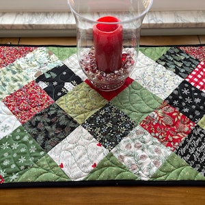 May include: A quilted table runner with a black border and a patchwork design featuring various holiday-themed fabrics in red, green, white, and black. The fabrics feature patterns of snowflakes, pine trees, houses, and text.