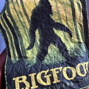 Puede incluir: Una manta acolchada negra y verde con una silueta de Bigfoot caminando por un bosque. El texto "Bigfoot The Missing Link" est&aacute; impreso en amarillo en la parte inferior de la manta.