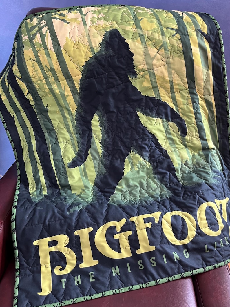 Puede incluir: Una manta acolchada negra y verde con una silueta de Bigfoot caminando por un bosque. El texto "Bigfoot The Missing Link" est&aacute; impreso en amarillo en la parte inferior de la manta.