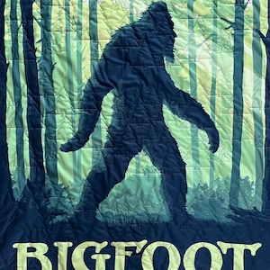 Puede incluir: Una tela acolchada azul oscuro con una silueta verde y blanca de Bigfoot caminando por un bosque. El texto "BIGFOOT THE MISSING LINK" est&aacute; impreso en amarillo en la parte inferior.