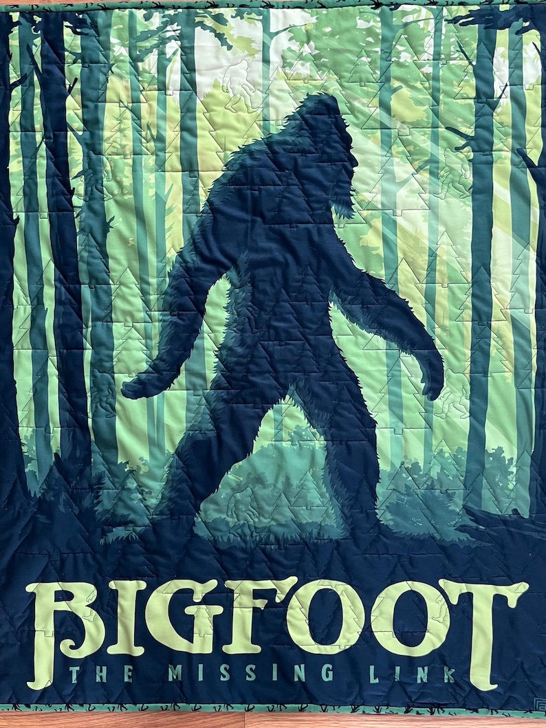 Puede incluir: Una tela acolchada azul oscuro con una silueta verde y blanca de Bigfoot caminando por un bosque. El texto "BIGFOOT THE MISSING LINK" est&aacute; impreso en amarillo en la parte inferior.