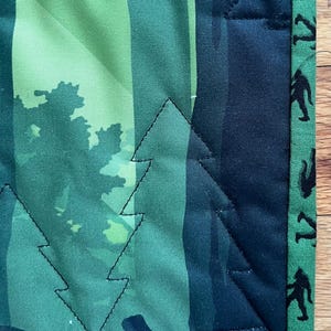 Puede incluir: Una colcha de tela verde y azul con un borde negro. La colcha presenta un patr&oacute;n de &aacute;rboles y un dise&ntilde;o repetitivo de figuras negras en el borde.