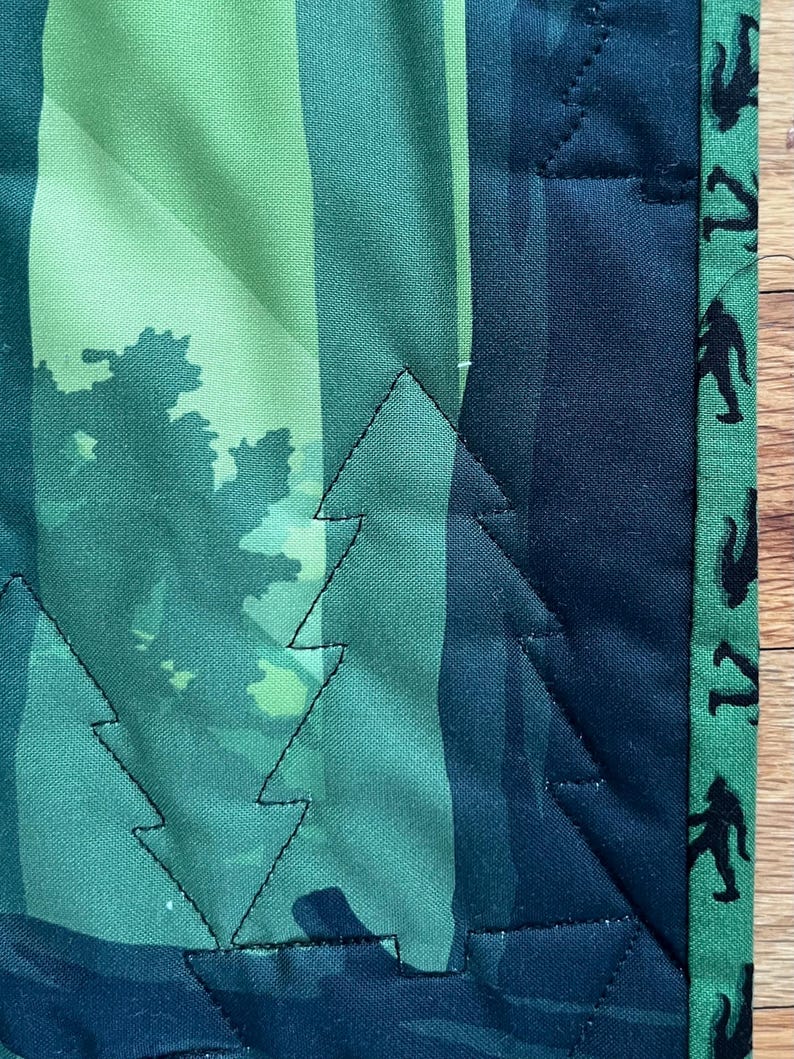 Puede incluir: Una colcha de tela verde y azul con un borde negro. La colcha presenta un patr&oacute;n de &aacute;rboles y un dise&ntilde;o repetitivo de figuras negras en el borde.