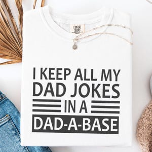 Op de afbeelding: Wit crewneck T-shirt met de zwarte tekst "I KEEP ALL MY DAD JOKES IN A DAD-A-BASE". Het shirt heeft een Comfort Colors label en een zilveren ketting met een kleine hanger.