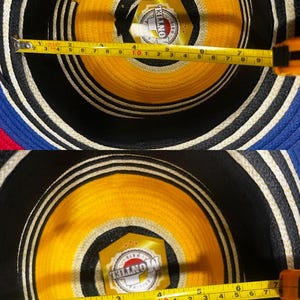 May include: A woven hat with concentric rings of blue, black, white, and yellow. A yellow label with the text MONTIEL HATS is visible inside. A yellow measuring tape shows the interior diameter is approximately 7.5 inches across the center.