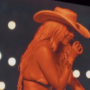 May include: A light-colored cowboy hat with a wide brim and a textured band, worn by an individual with long wavy hair. The individual is holding a black microphone near their mouth, set against a dark background with soft, out-of-focus circular lights.