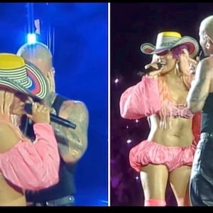 May include: A performer with long pink hair wears a pink two-piece outfit consisting of a long-sleeved crop top and puffy shorts, paired with a multi-colored cowboy hat featuring yellow, white, black, and red stripes, while holding a black microphone.